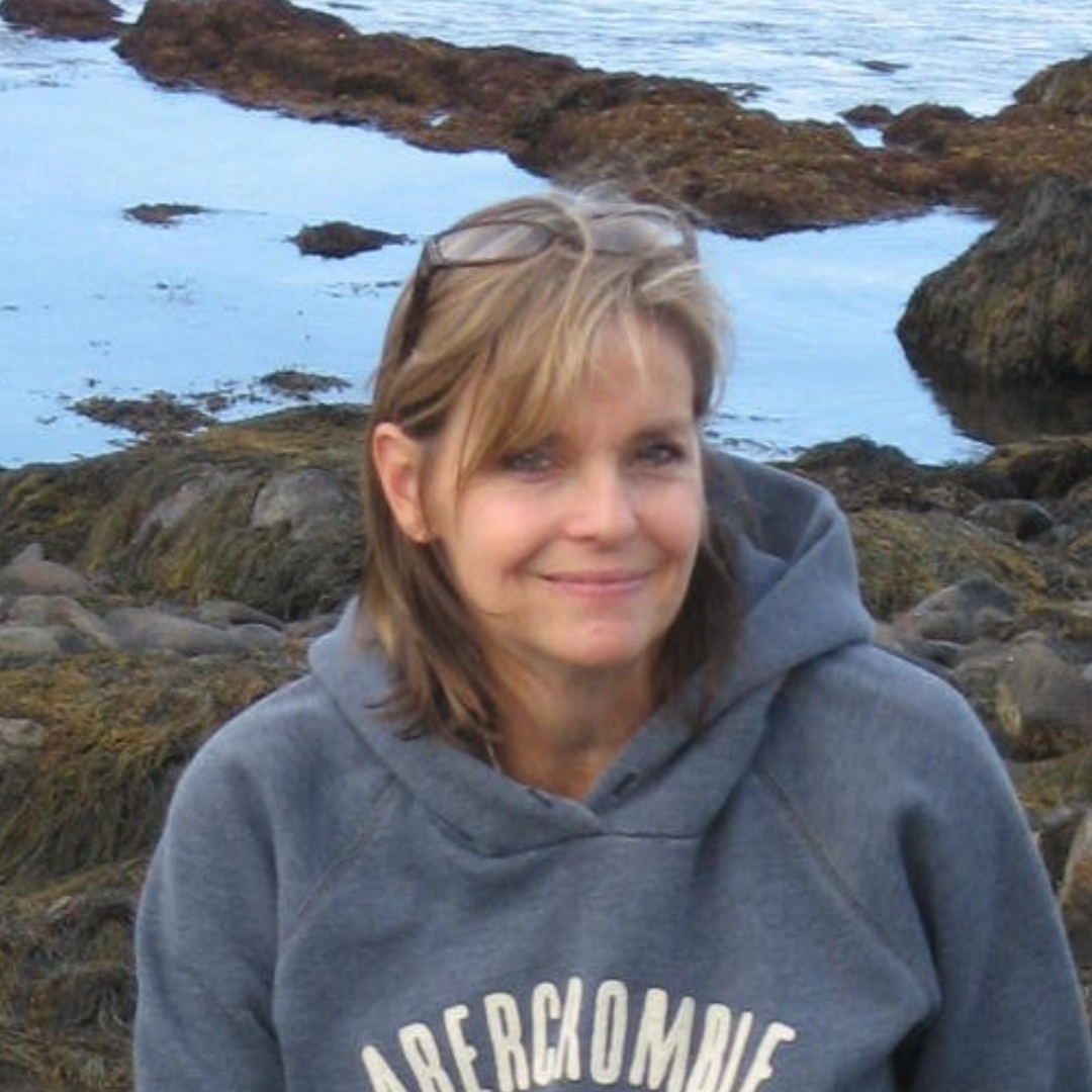 Jeanne, a woman smiling calmly at the camera in a cozy hoodie with a scenic body of water behind her